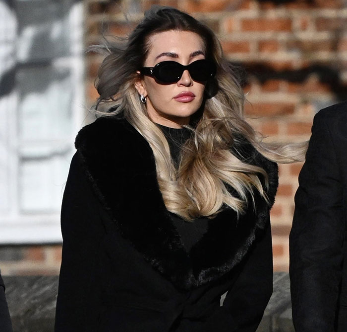 Woman in black coat and sunglasses, outdoors with windblown hair, looking pensive in front of a brick building.