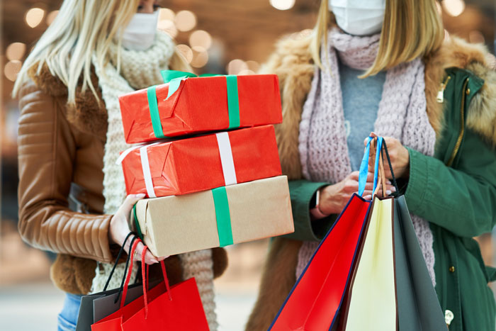 Women holiday shopping, holding gift boxes and shopping bags, wearing masks and scarves indoors.