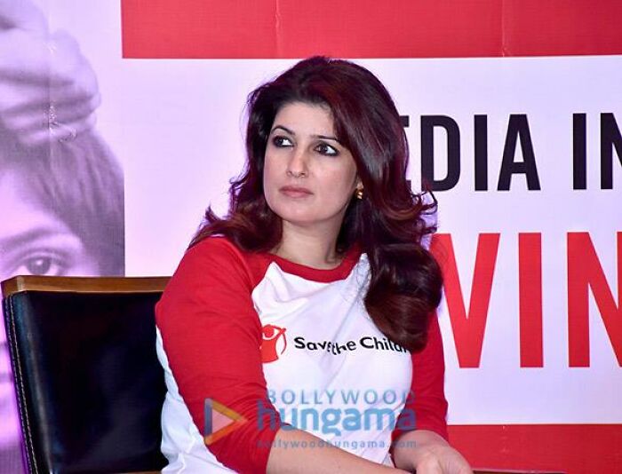 Celebrity in a red and white shirt at a public event, seated against a promotional backdrop.