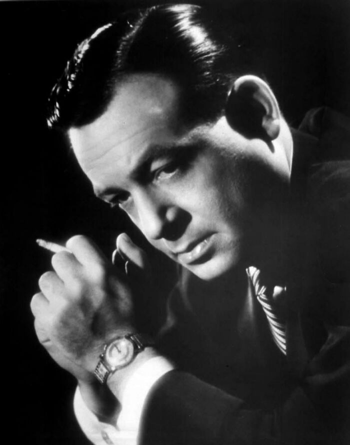 Black and white portrait of a man in a suit, holding a cigarette, representing a celebrity.