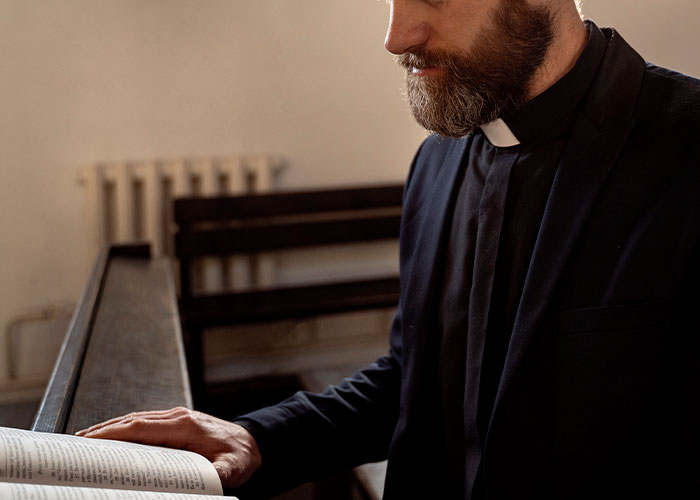 A bearded man in a clerical collar reads a book in a dimly lit room, illustrating intuition and gut feelings.