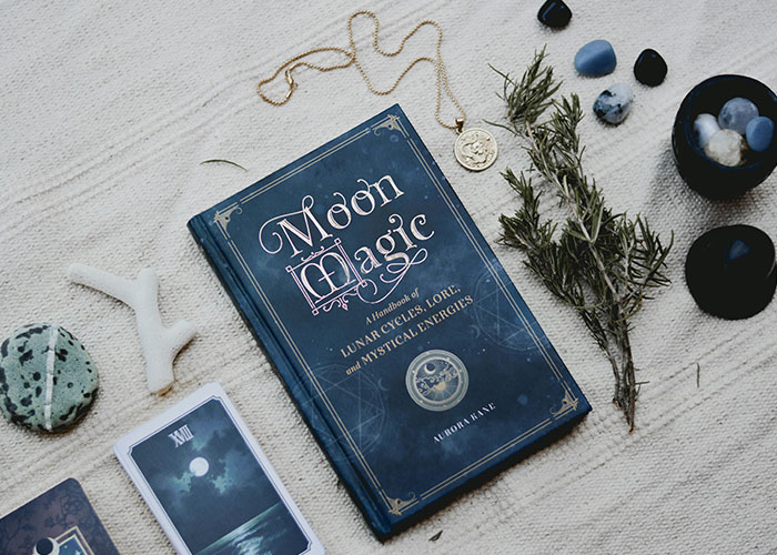 A book titled "Moon Magic" on a cloth with tarot cards, stones, herbs, and jewelry, suggesting mystical themes.