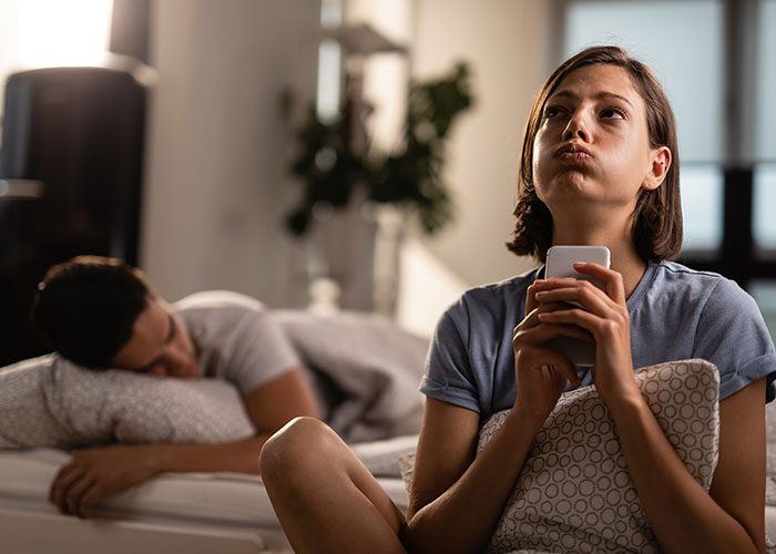 Woman with a concerned expression holding a phone, feeling uneasy while sitting on a bed.