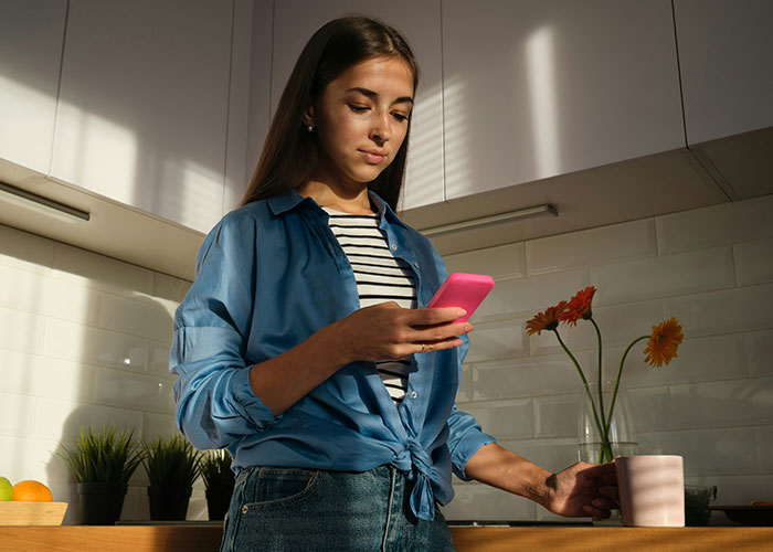 Woman in kitchen, holding pink phone, illustrating clever payback to boss on days off.