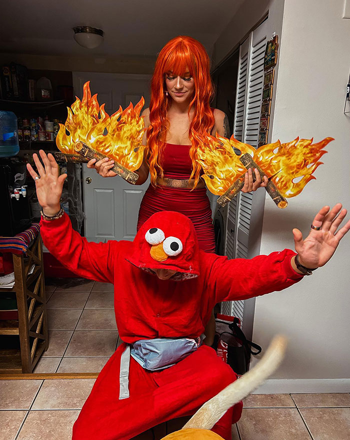 A person dressed as Elmo raises their arms in an "Elmo fire meme" pose, while another person with red hair holds fake flames behind them.