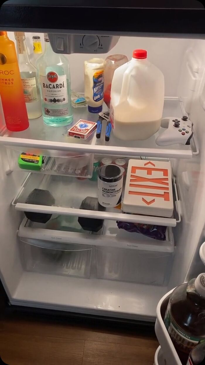 Chaotic dude fridge with drinks, milk, mayonnaise, a game controller, and random items on shelves.