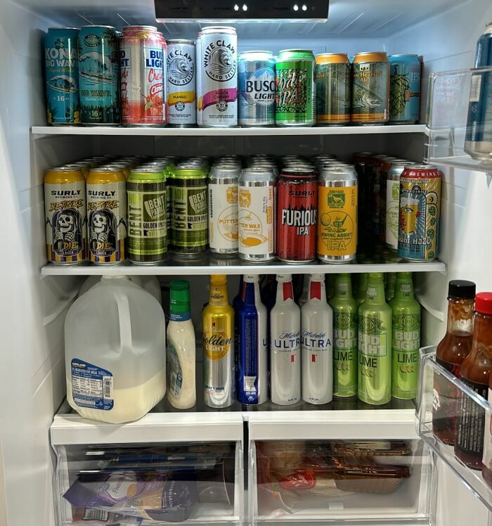 Open fridge filled with a variety of canned beers and beverages, milk, and condiments, showcasing a typical 'dude fridge'.