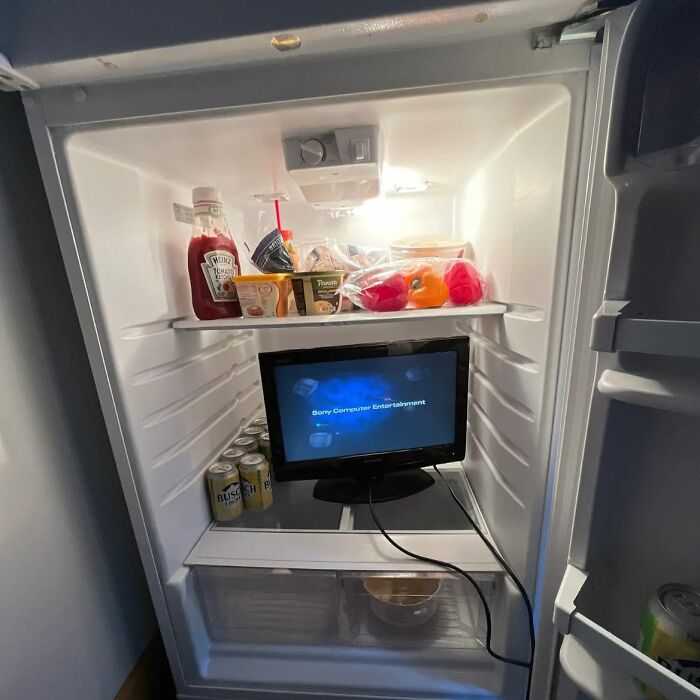 Open fridge with ketchup, drinks, and a small screen inside, showcasing a chaotic 'dude fridge' setup.