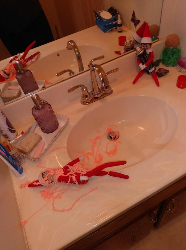 "Elf On The Shelf idea with one elf holding a can of silly string and another elf covered in pink silly string on the bathroom counter."