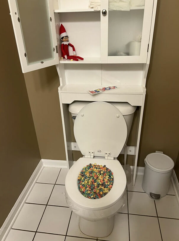 "Elf On The Shelf idea with an elf sitting above a toilet filled with colorful cereal, creating a humorous scene in the bathroom."