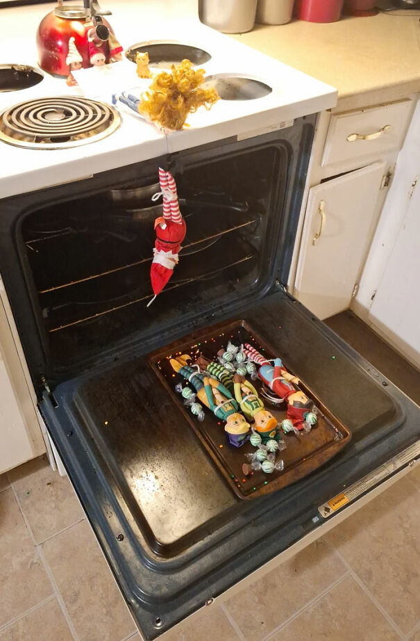 "Elf On The Shelf idea featuring an elf dangling over an open oven with toy elves arranged on a baking tray, creating a playful mischief scene."