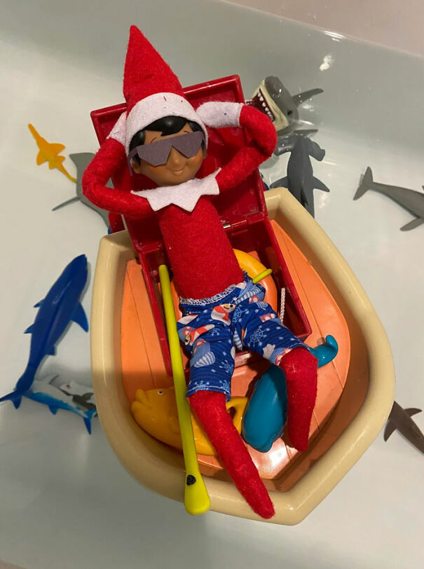 "Elf On The Shelf idea featuring an elf relaxing in a toy boat wearing sunglasses and swim trunks, surrounded by toy sharks in a bathtub."