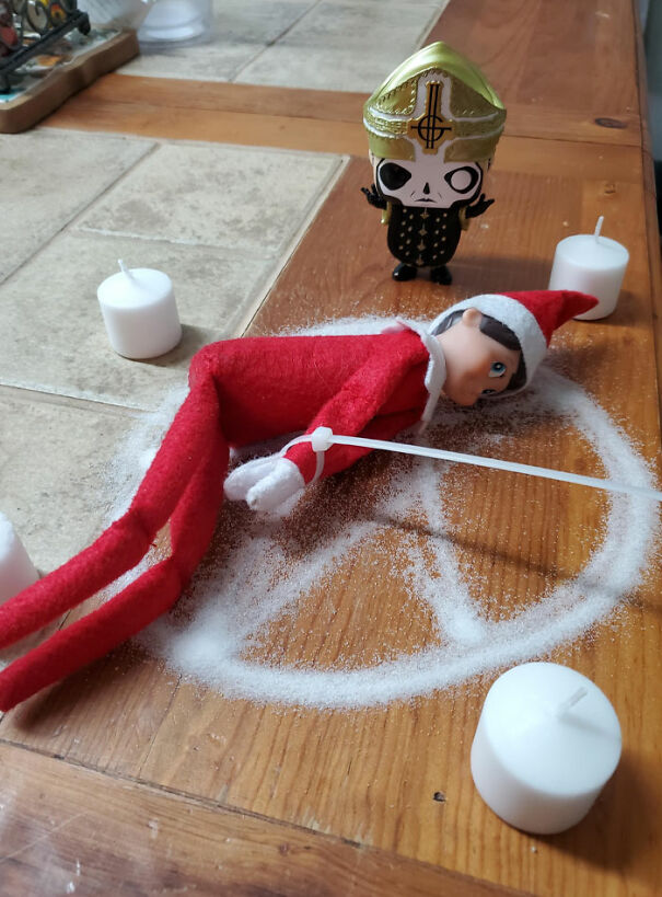 "Elf On The Shelf idea with the elf in a 'sacrificial ritual' setup, surrounded by candles and lying within a salt circle, with a small figure standing nearby."