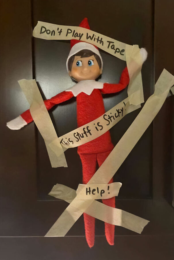 "Elf On The Shelf idea with the elf taped to a door with masking tape labeled 'Don't Play With Tape,' 'This Stuff is Sticky,' and 'Help!'"