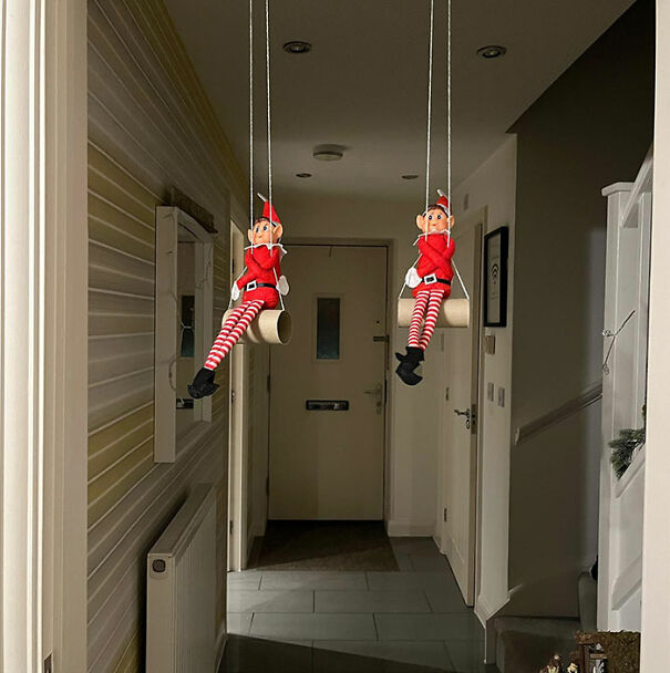 "Elf On The Shelf idea with two elves sitting on swings made from toilet paper rolls, hanging from the ceiling in a hallway."