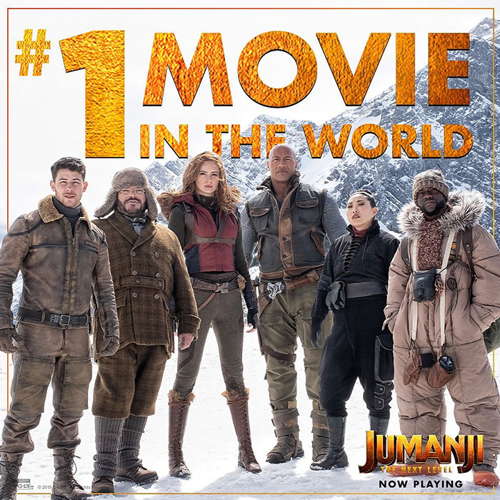 The cast of "Jumanji: The Next Level," including Kevin Hart in a fur-trimmed jacket, stands in a snowy mountain setting with bold text reading "#1 Movie in the World."