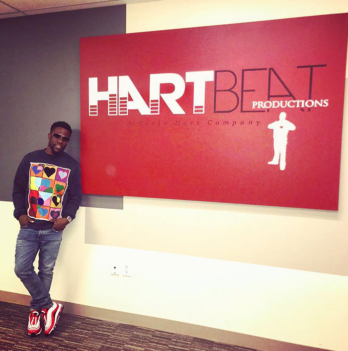 Kevin Hart leans casually against a wall with a red sign that reads "HartBeat Productions, A Kevin Hart Company," wearing a colorful heart-patterned sweater.