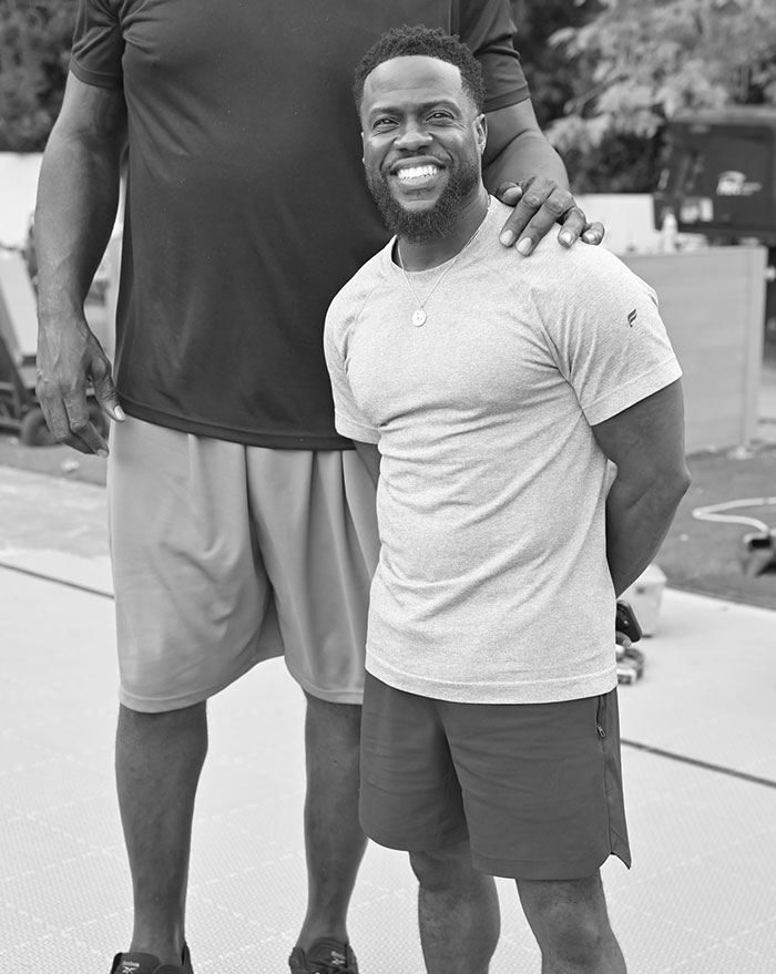 Kevin Hart smiles brightly in a casual outfit, standing next to a much taller individual whose hand rests on his shoulder, showcasing their height contrast.