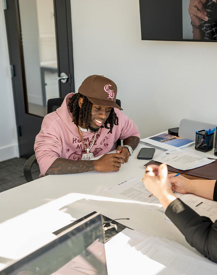 Kai Cenat wearing a pink hoodie and brown hat, sitting at a desk reviewing documents with a smile, representing professional growth.