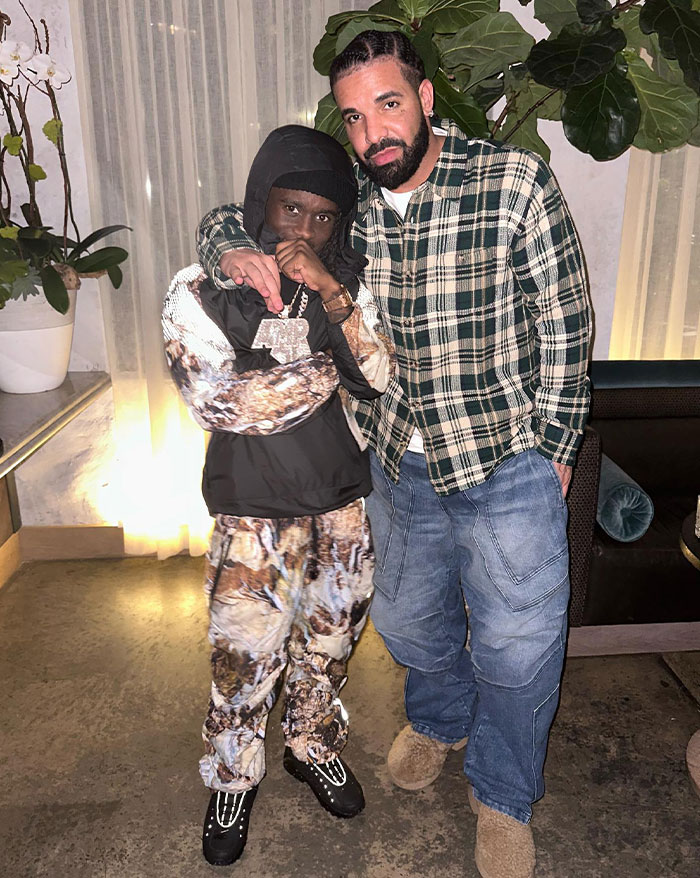 Kai Cenat posing with Drake, wearing a plaid shirt, standing in a well-lit space with plants, showcasing camaraderie and style.