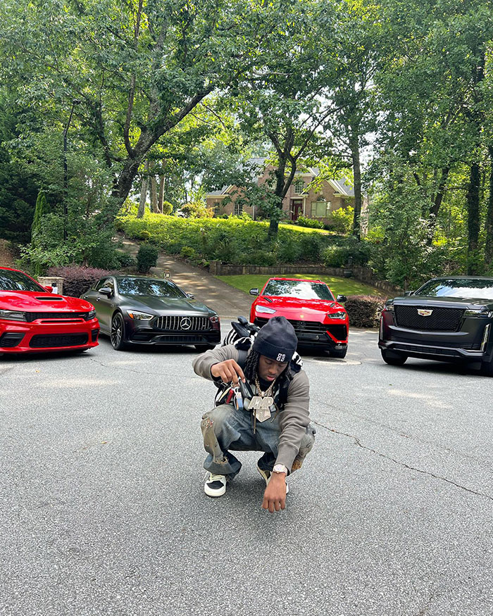 Kai Cenat crouching on a driveway with a set of car keys, surrounded by luxury cars, showcasing an opulent lifestyle amidst lush greenery.