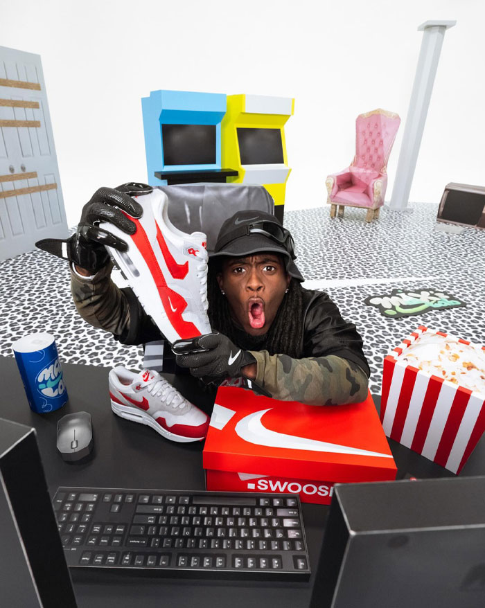 Kai Cenat excitedly holding a red-and-white Nike sneaker, surrounded by a playful setup with leopard-pattern flooring, arcade machines, and popcorn.