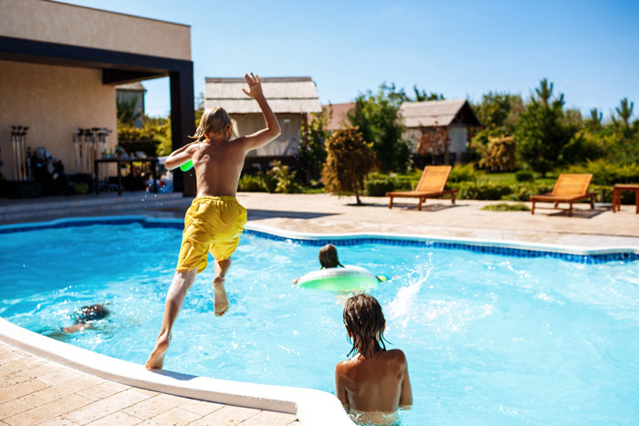 Kids playing alone in a pool, with one jumping in and others swimming under a clear blue sky.