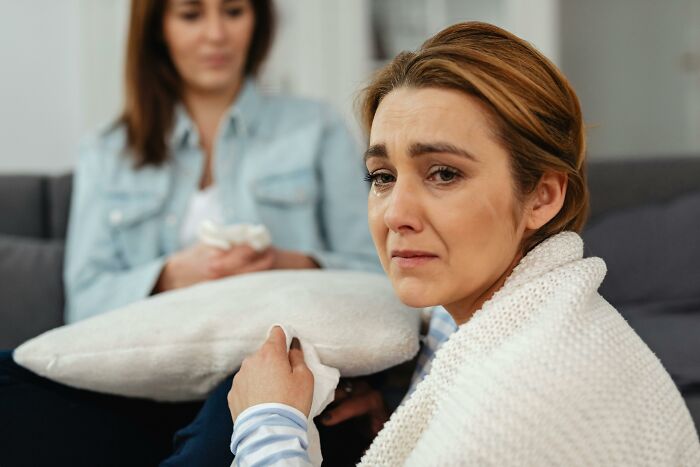 Grieving mother-in-law looking upset on a couch, while another woman listens in the background.