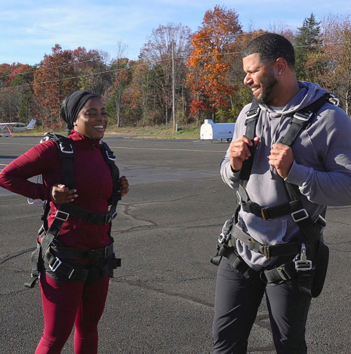 A couple wearing parachute harnesses, standing outside, smiling at each other on a sunny day.