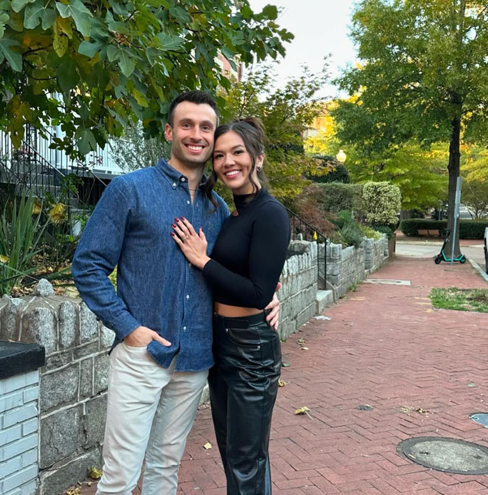 Couple from “Love Is Blind” smiling outdoors, embracing in casual attire by a brick walkway and lush greenery.