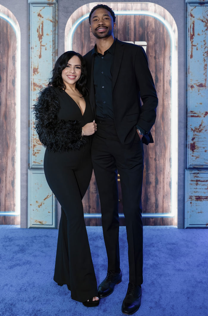 Couple from “Love Is Blind” in elegant black outfits, posing together on a blue carpet.