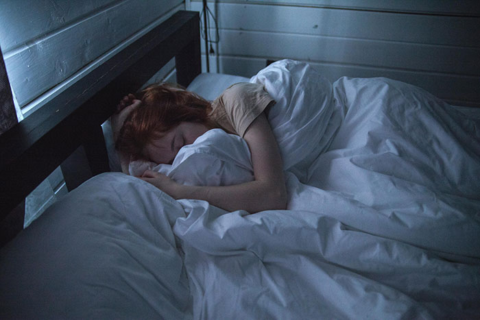 Woman sleeping in bed, unaware of perceived invasion, white sheets and dim lighting contribute to a tranquil setting.