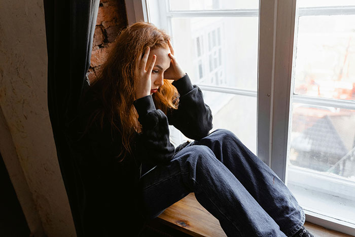 Young woman sitting by a window, holding her head in regret over revealing a secret.