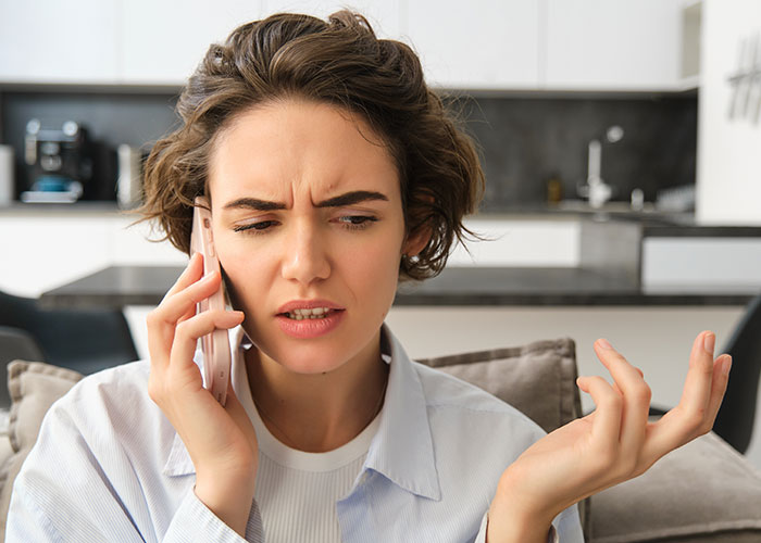 Woman on phone, looking puzzled, in a modern kitchen, discussing new neighbors' expectations in a non-Hallmark neighborhood.