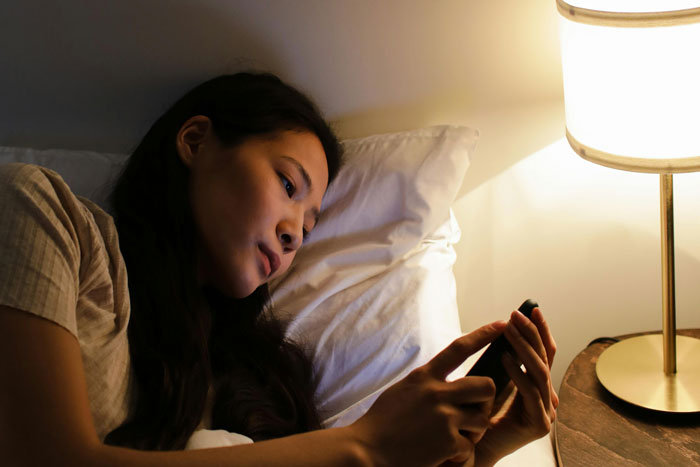 A person lying in bed, looking at their phone, illuminated by a bedside lamp, conveying a sense of being stalked.