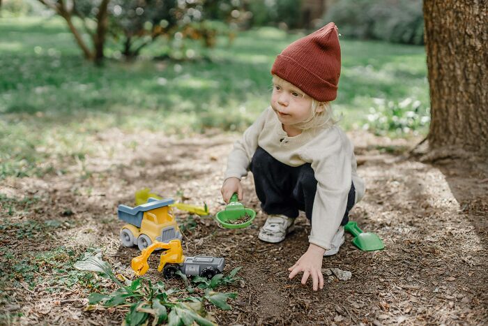 Child in a red beanie playing with toy trucks and a shovel outdoors, exploring the soil.