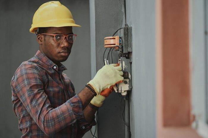 Worker in a yellow hard hat and gloves fixing wires outdoors, reminiscent of a funny joke you still think about to this day.