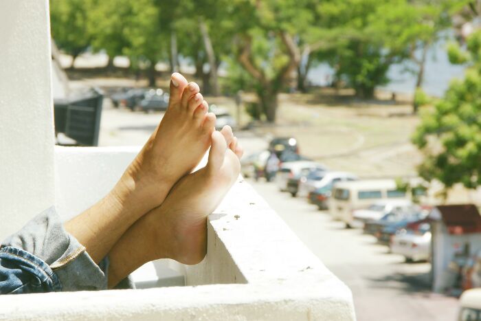 Bare feet resting on a balcony railing overlooking a street with cars and trees, symbolizing altered bodies.