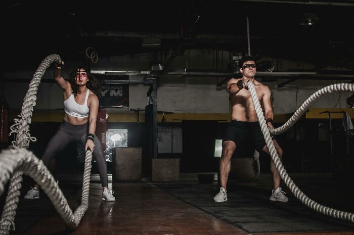 Two people exercising with battle ropes in a gym, working out intensely under dim lighting.