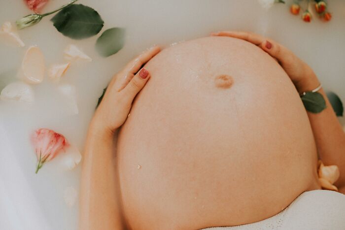 Pregnant belly in a milky bath surrounded by flowers, symbolizing altered bodies unknowingly.