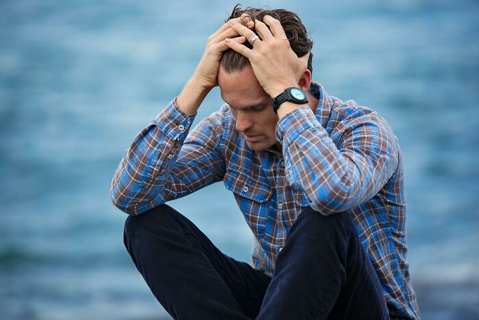 Man in plaid shirt sitting by the water, holding his head in distress, reflecting on how bodies are unknowingly altered.