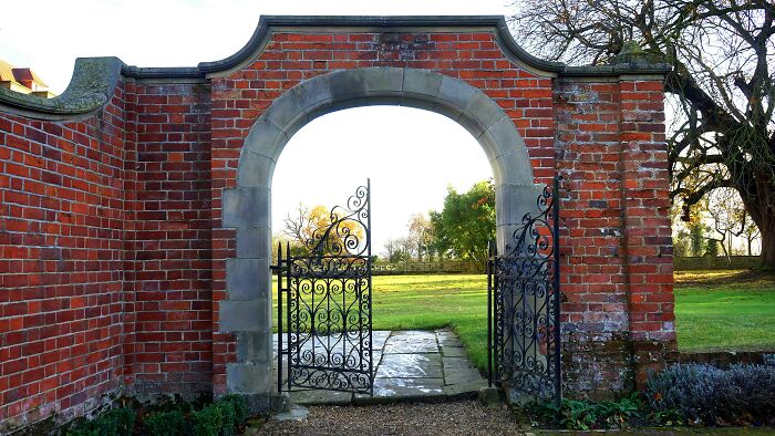 Ornate wrought iron gate in a brick wall, revealing a lush, expansive garden, suggesting luxury and wealth.