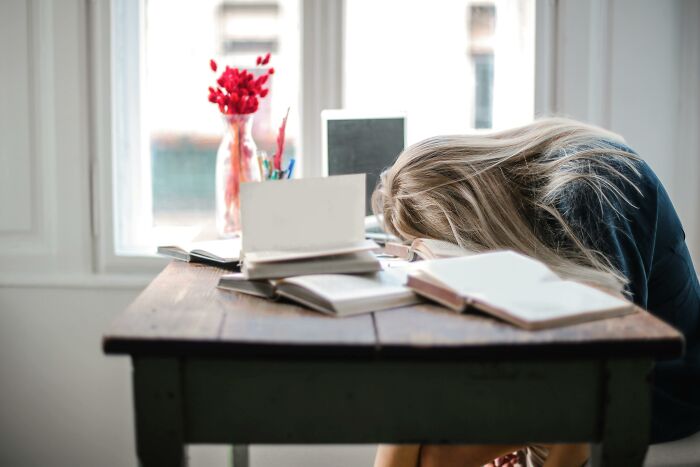 A person with head on table, surrounded by books, looking exhausted by the window; represents unknowingly altered bodies.