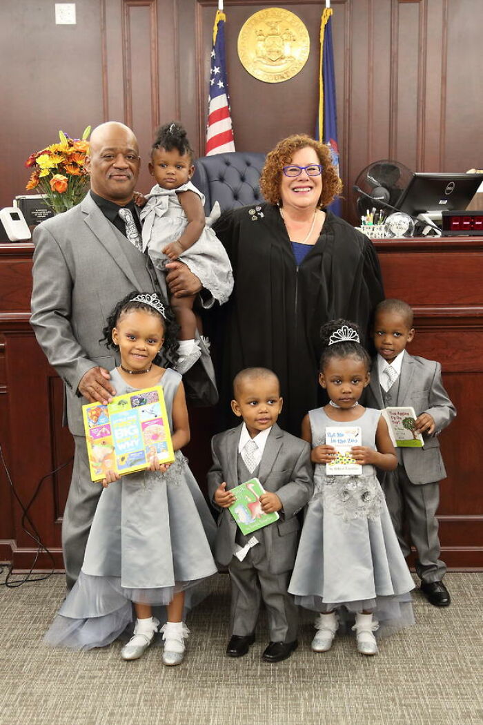 Family celebrates adoption with judge in court, children holding books and smiling, capturing a wholesome adoption moment.