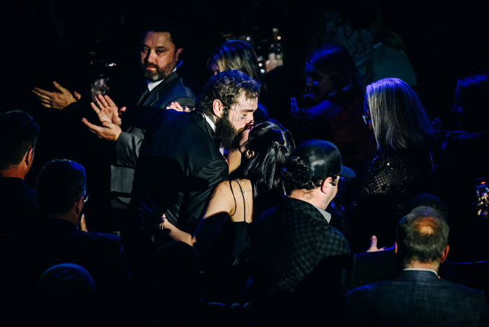 A candid moment of Post Malone embracing his fiancée in a dimly lit event setting, surrounded by a seated crowd in formal attire.