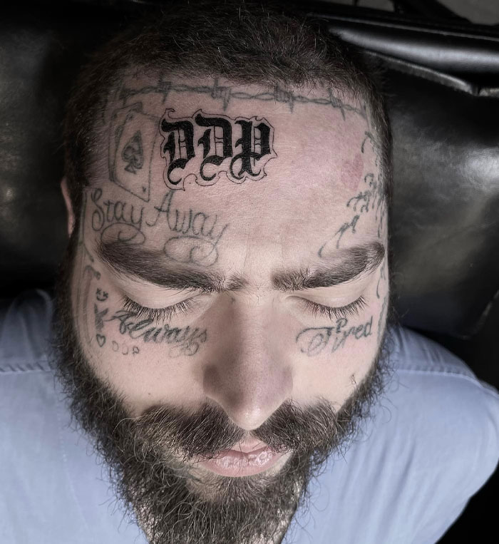 A close-up of Post Malone with his eyes closed, showcasing his facial tattoos prominently, including "Stay Away" and "DDP" on his forehead.