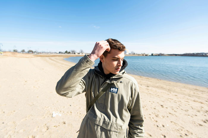 Teen standing on a windy beach, adjusting his jacket, thoughtful expression under a clear blue sky.