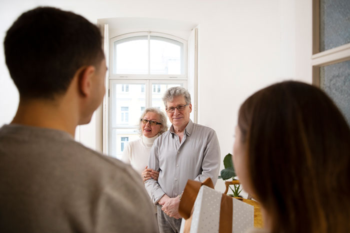 Teen with extended family in a living room, facing grandparents, holding gift bags.