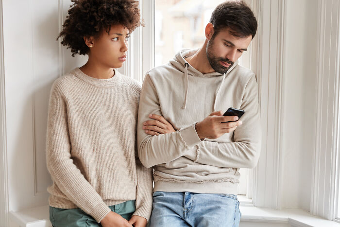 Couple in casual clothing sitting by a window, looking distant, person using phone; netizens share moments of doubt in relationships.