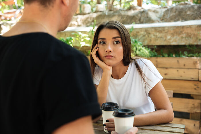 Woman with a doubtful expression holding coffee, listening to a man, illustrating a moment of uncertainty in a relationship.